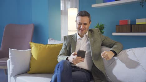 Casual-dressed-carefree-man-uses-smartphone-indoors-on-sofa-at-home.